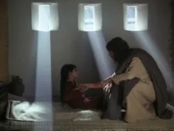 A man comforts a child sitting on a bed with sunlight streaming through three windows.